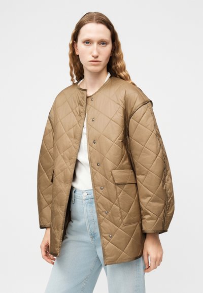 Γυναίκα που φορά μια μπεζ καπιτονέ μπουφάν με κουμπιά τύπου σναπ, εμπρόσθιες τσέπες, λευκό πουκάμισο και ανοιχτό μπλε τζιν, σε απλό φόντο.