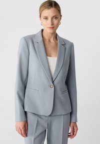 Blazer grigio chiaro su misura con un singolo bottone dorato, collo a rever e due tasche frontali. Tessuto liscio, vestibilità strutturata.