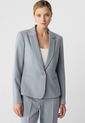 Blazer grigio chiaro su misura con un singolo bottone dorato, collo a rever e due tasche frontali. Tessuto liscio, vestibilità strutturata.