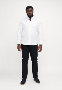 Camicia bianca con bottoni, maniche lunghe, pantaloni neri e sneaker nere, in piedi contro uno sfondo semplice.