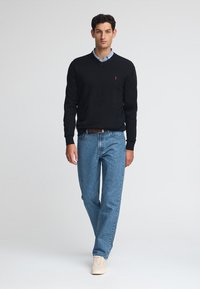 Maglione nero a v-neck, camicia blu sotto, jeans azzurri chiari, sneakers crema; design semplice, senza motivi, silhouette aderente.