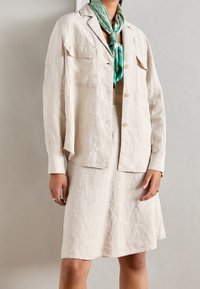 Giacca in lino beige con chiusura a bottoni e due tasche sul petto, abbinata a una gonna coordinata di lunghezza al ginocchio. Sciarpa verde a motivo annodata al collo.