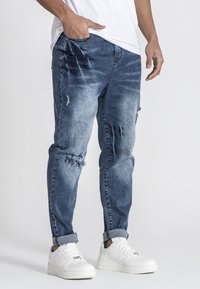 Jeans de mezclilla en azul con un efecto deslavado y detalles desgastados. Con puños enrollados y bolsillos frontales, combinados con zapatillas blancas.