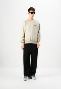 Sweatshirt beige avec des poignets et un ourlet côtelés, orné d'un petit logo texte. Associé à un pantalon noir surdimensionné et des baskets à accents bleus.