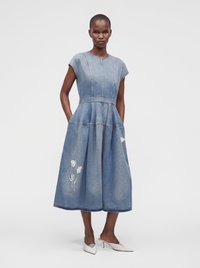 Γυναίκα με μπλε denim midi φόρεμα χωρίς μανίκια με κεντήματα λουλουδιών και τσέπες, συνδυασμένο με λευκές μυτερές γόβες.