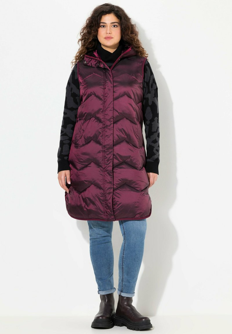 Giubbotto lungo e voluminoso di un borgogna scuro, con cappuccio e motivo a zig-zag. Abbinato a un top nero con fantasia e jeans blu, completato da stivali scuri.
