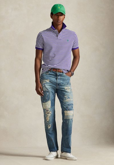 Άνδρας που φοράει πράσινο καπέλο, μωβ ριγέ πόλο πουκάμισο, τζιν με σκισίματα patchwork, καφέ ζώνη, ρολόι και λευκά αθλητικά παπούτσια, στέκεται μπροστά σε απλό φόντο.