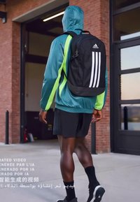 Person wearing teal-green hooded jacket and black shorts walks outdoors carrying a black Adidas backpack with white stripes.