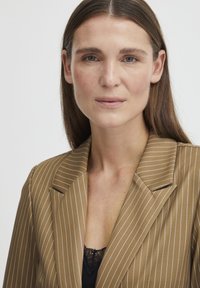 Pruun ribaülik, mis on valmistatud siledast kangast, millel on soonikuga krae ja üks disksed tasku detail, kihtide kaupa musta pitsist topi peal.