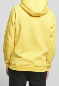 Osoba v jednobarevném žlutém mikině a černých kalhotách, vyfotografovaná zezadu na neutrálním pozadí.