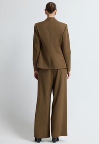 Ruskea räätälöity bleiseri yhdessä vastaavien leveälahkeisten housujen kanssa. Sileä kangas, rakenteelliset olkapäät ja minimaalinen yksityiskohtaisuus. Tarkoitettu ammattimaisen ulkoasun saavuttamiseksi.