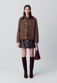 Giacca in misto lana marrone con dettagli dorati, due tasche frontali; abbinata a una gonna di pelle e stivali alti fino al ginocchio in suede marrone. Inclusa una borsa color marsala.