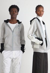 Felpe grigie con zip e cappuccio, con accenti neri e maniche bianche; materiale morbido, vestibilità comoda; indossate sopra completi bianchi e neri.