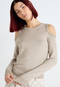 Maglione beige a maglia con design a spalle scoperte, impreziosito da borchie argentate sulle cuciture delle spalle, con maniche lunghe e polsini a costine.