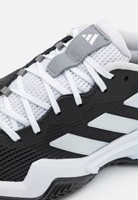Chaussure de sport noire et blanche avec une tige en maille, trois rayures argentées, semelle en caoutchouc et accents texturés. Les lacets sont blancs et la languette est rembourrée.