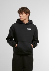 Black hooded sweatshirt made of soft fabric, featuring a front pocket and white text printing on the chest. Loose fit and casual design.