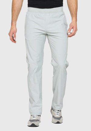 Uomo con pantaloni casual grigio chiaro e sneakers grigie e bianche, in piedi su uno sfondo chiaro e semplice.