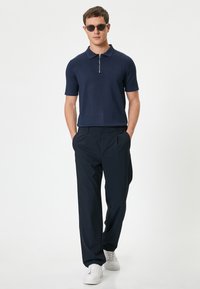 Mörkblå kortärmad polo med dragkedja i kragen, ihopparad med skräddarsydda mörkblå byxor och vita sneakers. Modellen bär runda solglasögon.