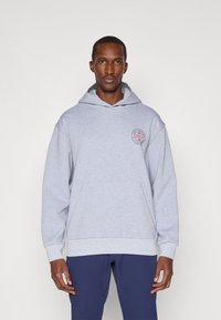 Grå hettegenser med lomme foran, ribbede mansjetter og et sirkulært logo på venstre brystområde. Glatt tekstur og avslappet passform.