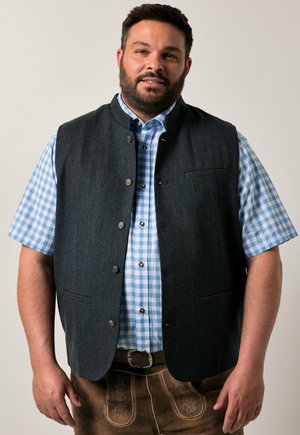 Donkergrijze wollen vest met een ronde halslijn en een knopenlijst, gedragen over een lichtblauw en wit geruit overhemd. Bruine lederhosen met decoratieve stiksels.