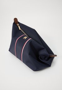Marinblå stoffpose med et gulllogo, en skrå glidelås og rød-hvite striper. Myk tekstur med en krøllet design.