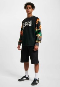 Black long-sleeve shirt featuring camouflage-patterned sleeves, gray cuffs, and white graphic text. Paired with knee-length black shorts and sneakers.