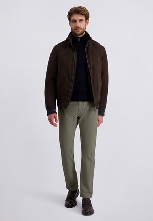 Mann trägt eine dunkelbraune Zip-Jacke, einen schwarzen Rollkragenpullover, olivgrüne Hosen und dunkelbraune Schuhe und steht vor einem weißen Hintergrund.