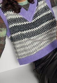Gestrickte ärmellose Weste in schwarz-weißer Zickzackmuster mit lila V-Ausschnitt und Saum. Strukturierter Stoff; verkürztes Design, kombiniert mit einem Lederoutfit.