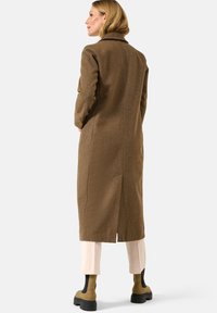 Bruine, lange wollen jas met een kraag, split aan de achterkant en een getailleerd ontwerp. Gecombineerd met lichte pantalons en beige enkellaarsjes. Gladde textuur.