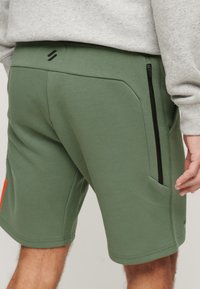 Pantaloni sportivi verdi caratterizzati da una texture liscia, tasche laterali e una zip nera, insieme a un logo sulla parte posteriore della vita.