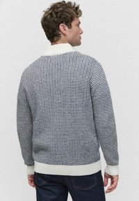 Man som står med ryggen mot kameran iklädd en blå och vit texturerad stickad tröja med ribbat krage, ärmar och fåll, tillsammans med mörkblå jeans.