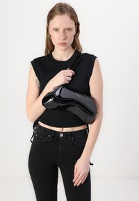 Crna lakirana torba na rame s zakrivljenim oblikom i sjajnom teksturom, držana blizu tijela, u kombinaciji s crnim kratkim topom i trapericama.