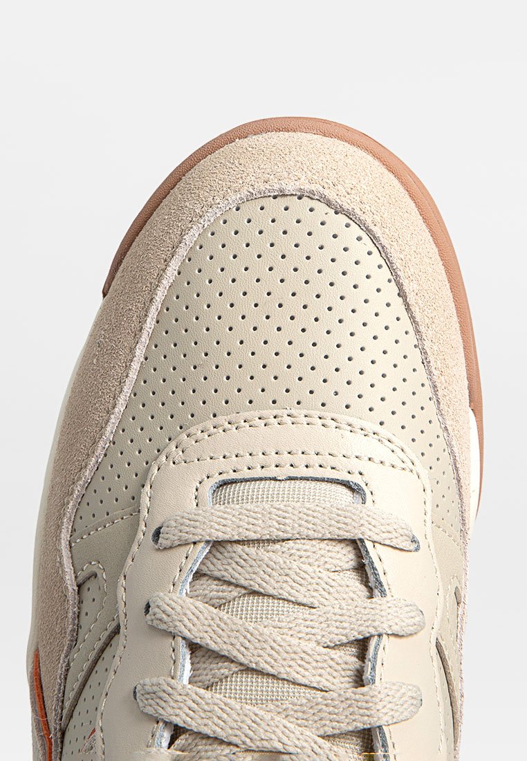 Sneaker beige con design perforato, dettagli in suede e lacci piatti. La suola in gomma presenta una rifinitura marrone. Superfici lisce e texture.