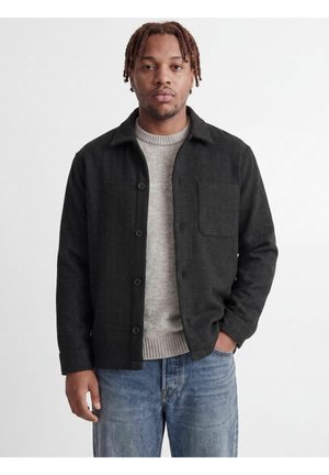 Mand med korte dreadlocks iført en sort skjortejakke over en grå sweater og blå jeans, der står mod en hvid baggrund.