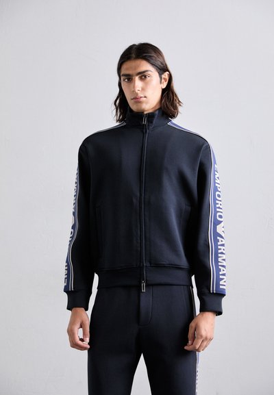 Granatowy rozpinany sweatshirt wykonany z miękkiego materiału, z wysokim kołnierzem, białymi paskami po bokach oraz logo "Emporio Armani" na rękawach.