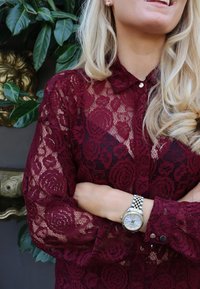 Bordó csipkeblúz virágmintákkal, galléros nyakkivágással és gombos záródással. Ezüst karóra mintás szíjjal.