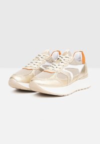Baskets métalliques dorées avec textures en mesh et cuir, dotées d'un logo blanc sur le côté et d'une languette arrière orange. Design léger avec une semelle épaisse blanche.