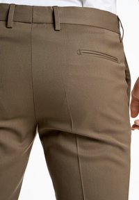 Pantaloni marroni su misura con una texture liscia, dotati di una tasca posteriore singola e passanti per cintura, mostrati da una vista leggermente inclinata.