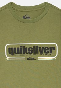 T-shirt verde oliva de algodão com decote redondo. Apresenta um grande logótipo preto "quiksilver" e linhas de destaque brancas. Design simples.