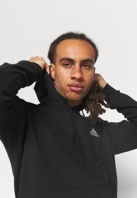 Sudadera negra de tela suave con un bolsillo frontal, cordones y un pequeño logo gris de Adidas en el pecho.