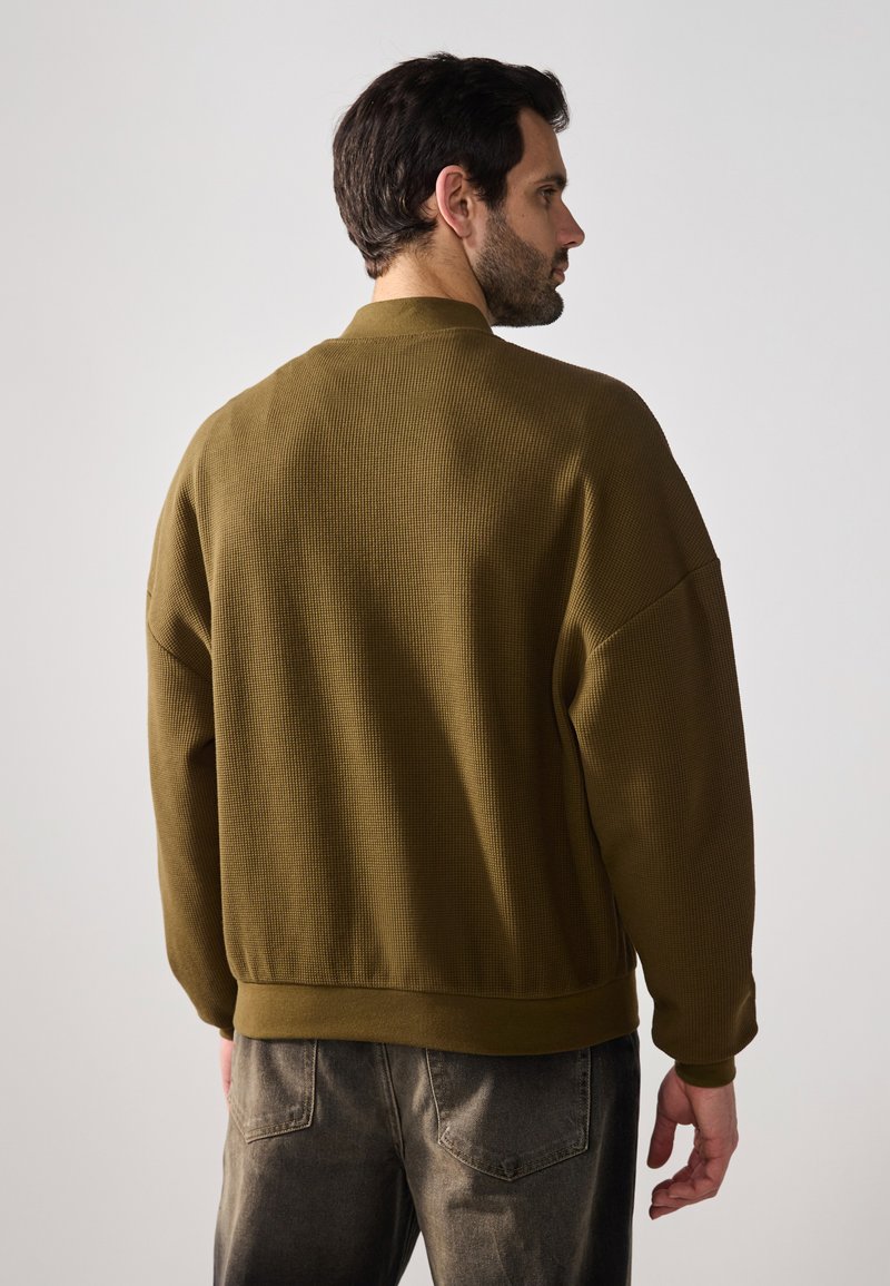 Olivgrüner, strukturierter, übergroßer Pullover mit geripptem Stoff und rundem Ausschnitt, kombiniert mit grauen Denim-Jeans.