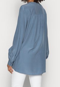 Blouse en soie bleu clair à manches longues, poignets boutonnés, coupe décontractée, plis au dos et ourlet arrondi. Texture lisse avec une légère brillance.