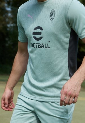 Mężczyzna ubrany w jasnozielony stroje piłkarski AC Milan i Puma stoi na zewnątrz na boisku sportowym.