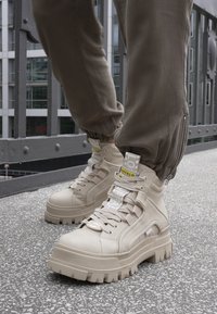 Des baskets montantes beiges avec une semelle épaisse, un dessus texturé, plusieurs sangles et des étiquettes de marque. Elles sont associées à un pantalon olive ajusté.