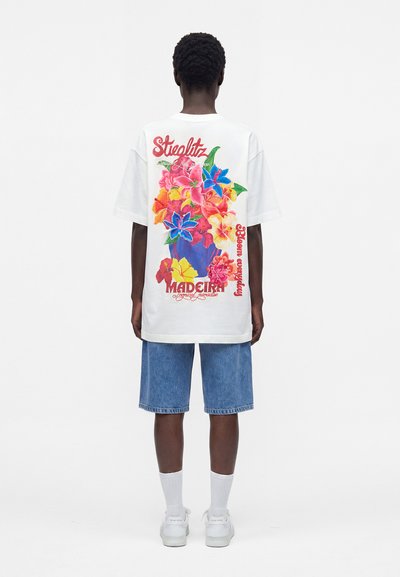 Άτομο που φοράει λευκό oversized T-shirt με πολύχρωμο floral print και κείμενο, σε συνδυασμό με τζιν σορτς μέχρι το γόνατο και λευκά αθλητικά παπούτσια.