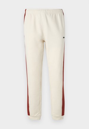Calças jogger creme com riscas laterais vermelhas, cintura elástica e bainhas com punhos. Feitas de uma mistura de algodão suave com detalhe do logo Lacoste.