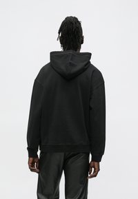 Persona ar dredveidiem, kas valkā melnu kapuci un melnas bikses, stāvot ar muguru pret vienkāršu pelēku fonu.