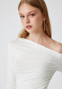 Witte, aansluitende off-shoulder top van een zachte, gladde stof met geplooide details aan één kant, gedragen met grote gouden creolen.