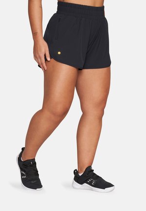 Atletyczna osoba ubrana w krótkie czarne spodenki z gumką w talii i kieszenią na zamek oraz czarne buty treningowe z białą podeszwą, stojąca na białym tle.