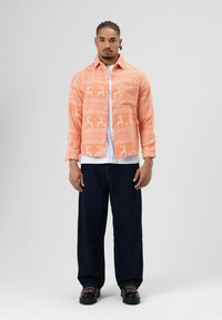 Oranje overhemd met patroon en hertmotieven, knoopsluiting en lange mouwen, gedragen over een wit T-shirt, gecombineerd met donkere spijkerbroeken en zwarte schoenen.